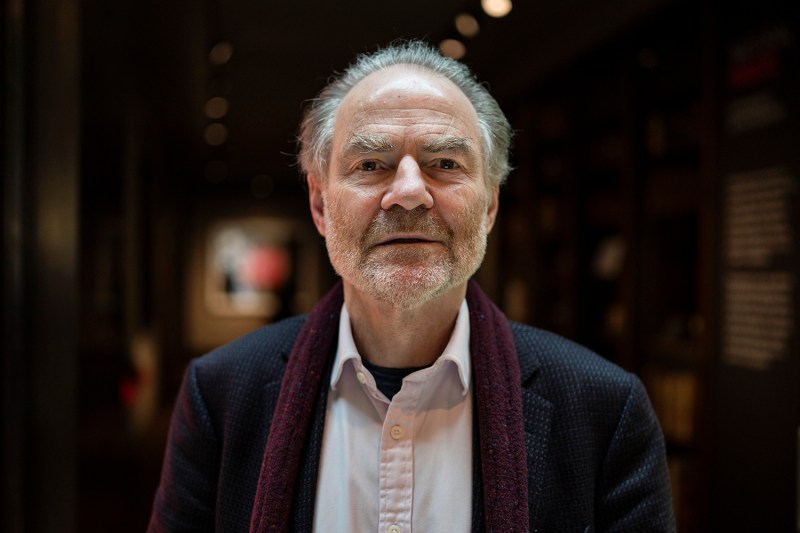 Timothy Garton Ash stares directly at the camera, his mouth slightly open. He is older than in the previous photos, hair and beard now entirely gray, and he wears a burgundy scarf with a dark gray blazer.