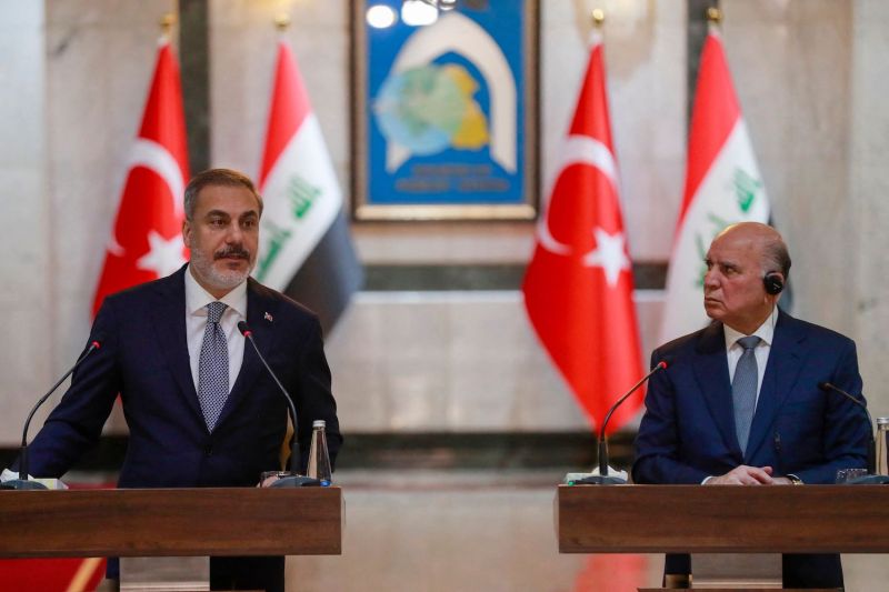 Turkish Foreign Minister Hakan Fidan speaks at a podium during a joint press conference. Next to him, Iraqi Foreign Minister Fuad Hussein stands behind his own podium and frowns as he watches Fidan. A marble wall is visible behind the men, and the flags of Iraq and Turkey stand in front of it.