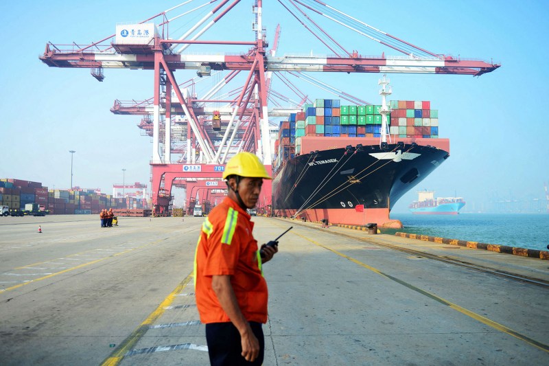 A worker wearing a hard hat and orange reflective shirt and holding a walkie talkie stands in front of a massive cargo ship docked under cranes as it's loaded at a port in Qingdao, China. Another ship floats on the water in the distance. Both are loaded with cargo containers.