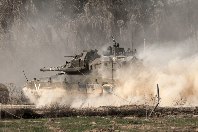 An Israeli army tank moves through a torn-up patch of grass in the northern part of the Gaza Strip. The tank kicks up dirt and dust from the ground, which billows in the air and partially obscures the tank, although a soldier is visible sitting on top of it with an automatic rifle.
