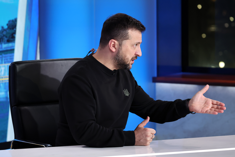 Ukrainian President Volodymyr Zelensky gestures with an open hand while speaking during a TV news interview. Zelensky is dressed in his typical black t-shirt and is seated at a desk in front of a bright blue wall.