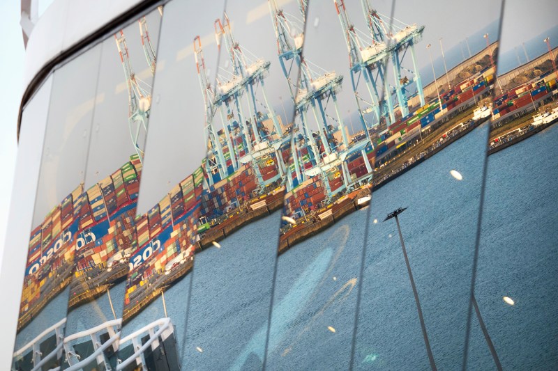 Reflections of the crains and cargo ships are visible in a reflection in the segmented panoramic windows of a moored ship. The ships are piled high with shipping containers in various colors, and a hazy sky is visible above them.