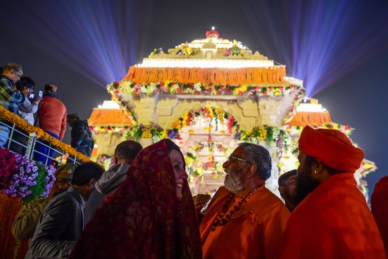 People in a crowd smile as they look up at the yellow exterior of the Ram Mandir temple, festooned with flowers, which looms above them as light beams illuminate the night sky.