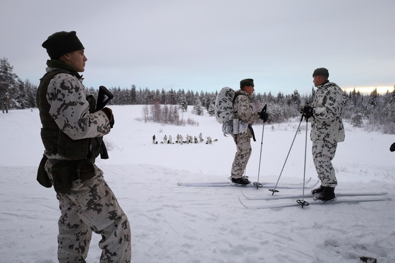 In a snowy landscape looking slightly downward from a hilltop, a side profile of a man holding a gun stands on the left of frame beside two other men on skis having a discussion. In the distant background, a group of military personnel stand in a large clearing with their equipment bags. Trees line the horizon and surround the environment.