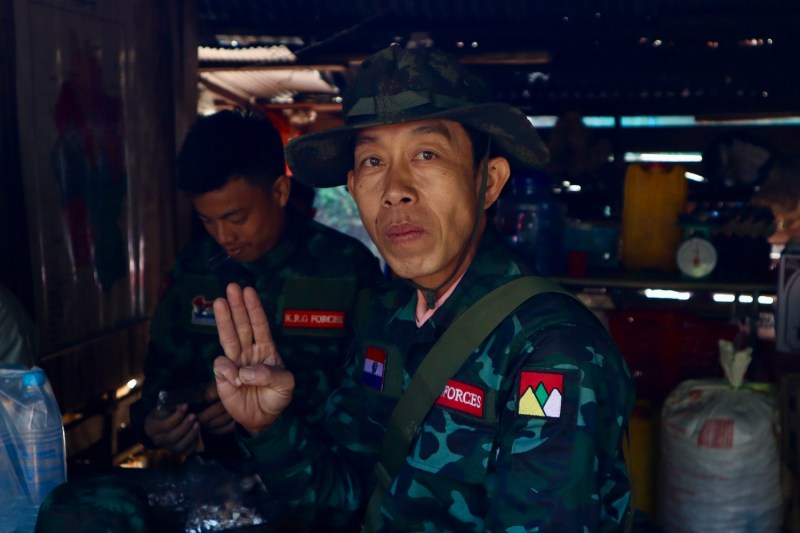 A Karenni Army soldier wearing a camouflage hat and uniform flashes the three-finger pro-democracy salute at a betel nut shop. Another soldier is seen behind him in the crowded space.