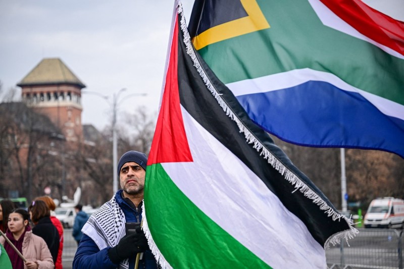 A man with a serious expression holds the Palestinian and South African flags as he stands at a demonstration in support for Palestinians in Bucharest, Romania, under a cloudy sky.