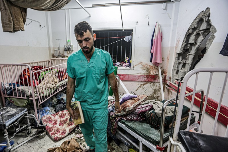 A man waring scrubs and holding a book walks through a damaged room at Nasser hospital in Khan Yunis in the southern Gaza Strip. A hole is seen in one wall and bedding, beds, and other equipment are tossed about the space.