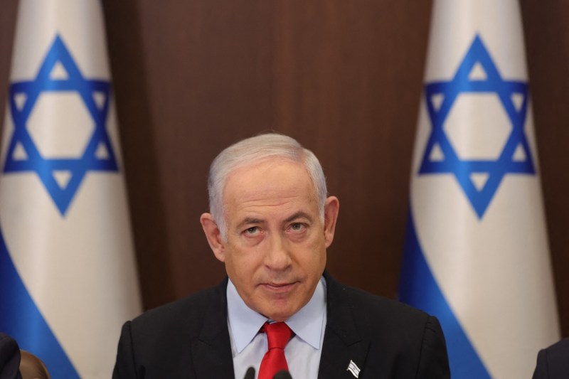 Benjamin Netanyahu stands between two Israeli flags in front of a microphone wearing a black suit with an Israel flag pin, blue shirt and red tie.