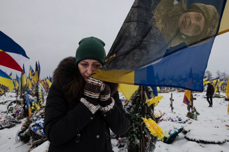 A woman wearing mittens, a coat, and a winter hat touches a flag with a soldier's face on it to her face. Behind her is a snowy graveyard scene with flowers and Ukrainian flags.