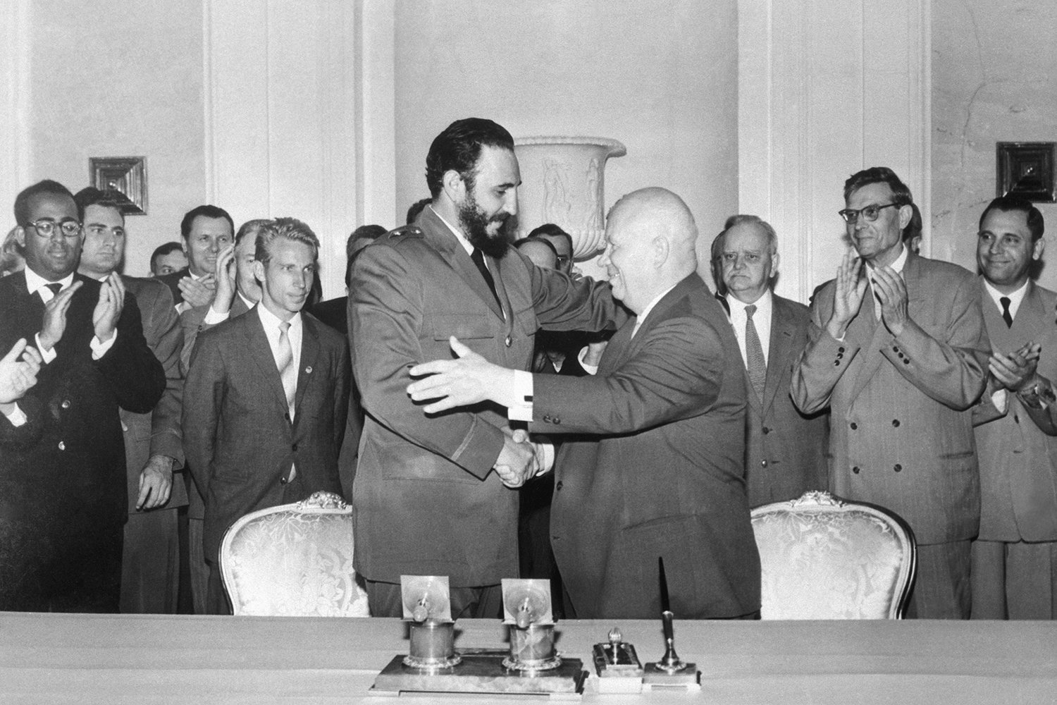 A historic black-and-white photo shows Fidel Castro and Nikita Khrushchev shaking hands in front of a group of men in suits during a meeting in Moscow. Castro is younger and nearly a head taller than Khruschev as he leans in to embrace him. Some of the men in the background clap as they watch.