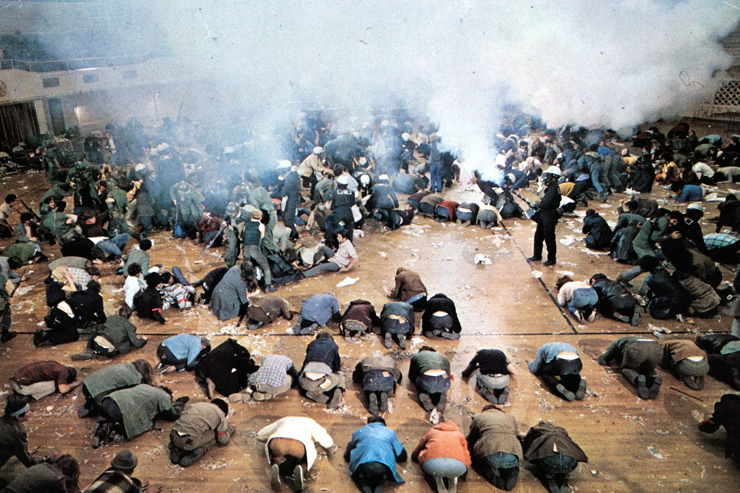 A crowd of students kneel on the ground with their faces pressed against it as they duck from clouds of tear gas in a film still from 1968.