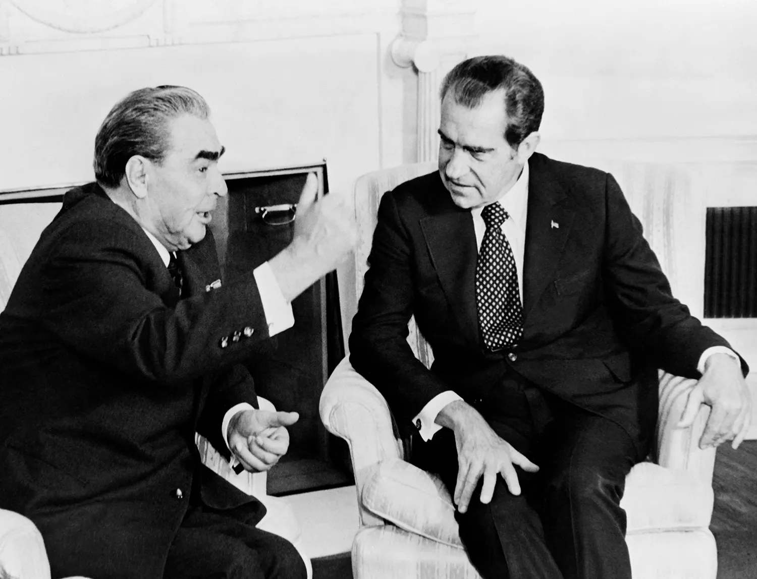 A historic black-and-white photo shows Leonid Brezhnev and Richard Nixon sitting and talking in the White House. Both men are midde-aged and wearing dark suits and ties. Brezhnev gestures with one hand in the air as he speaks, and Nixon leans in closer to listen with one hand balanced on his own knee.