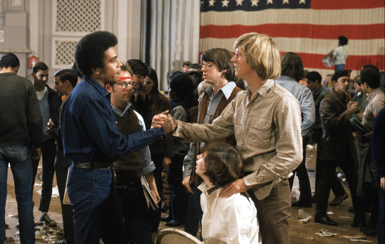 Two actors shake hands in a group, as they stand in a crowded room with a large American flag hanging against the far wall in this film still from 1968.