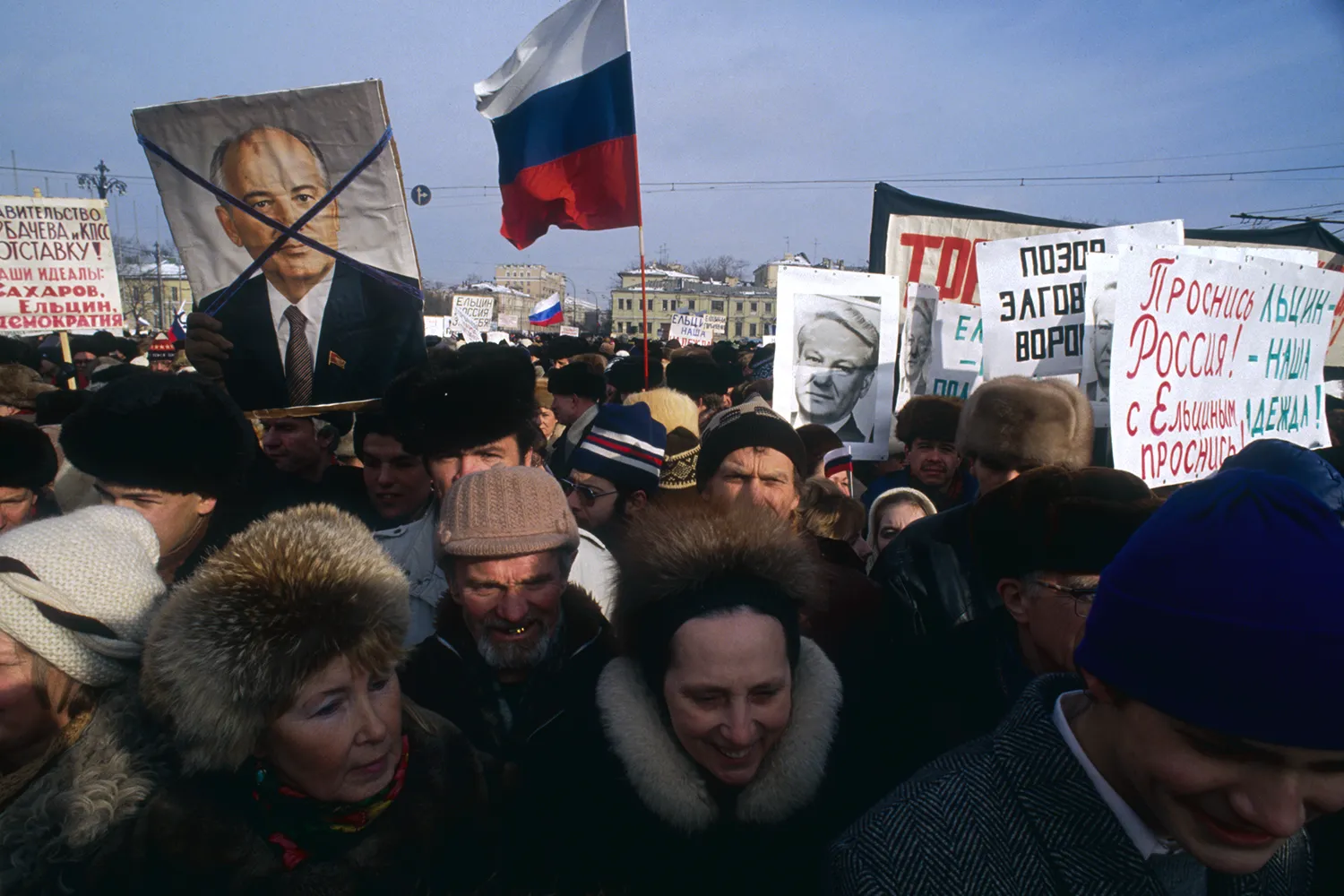 A crowd of demonstrators in Moscow protest Mikhail Gorbachev beneath a partly cloudy sky. Some hold Russian flags, while others wave picket signs or photos of Gorbachev with an X drawn over his face in marker.