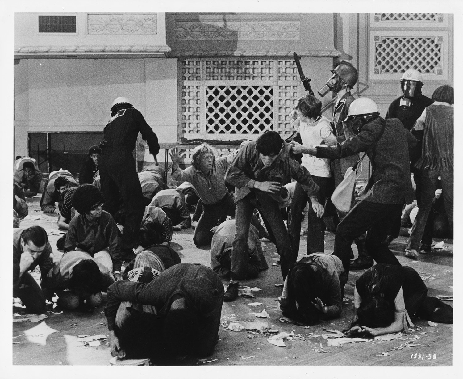 A group of students clash with police in a black and white film still from 1968. An officer in riot gear has his hand around one young woman's waist; others shield their heads with their hands while sheltering against the ground. A man at the middle of the frame has his hands raised up as he kneels.