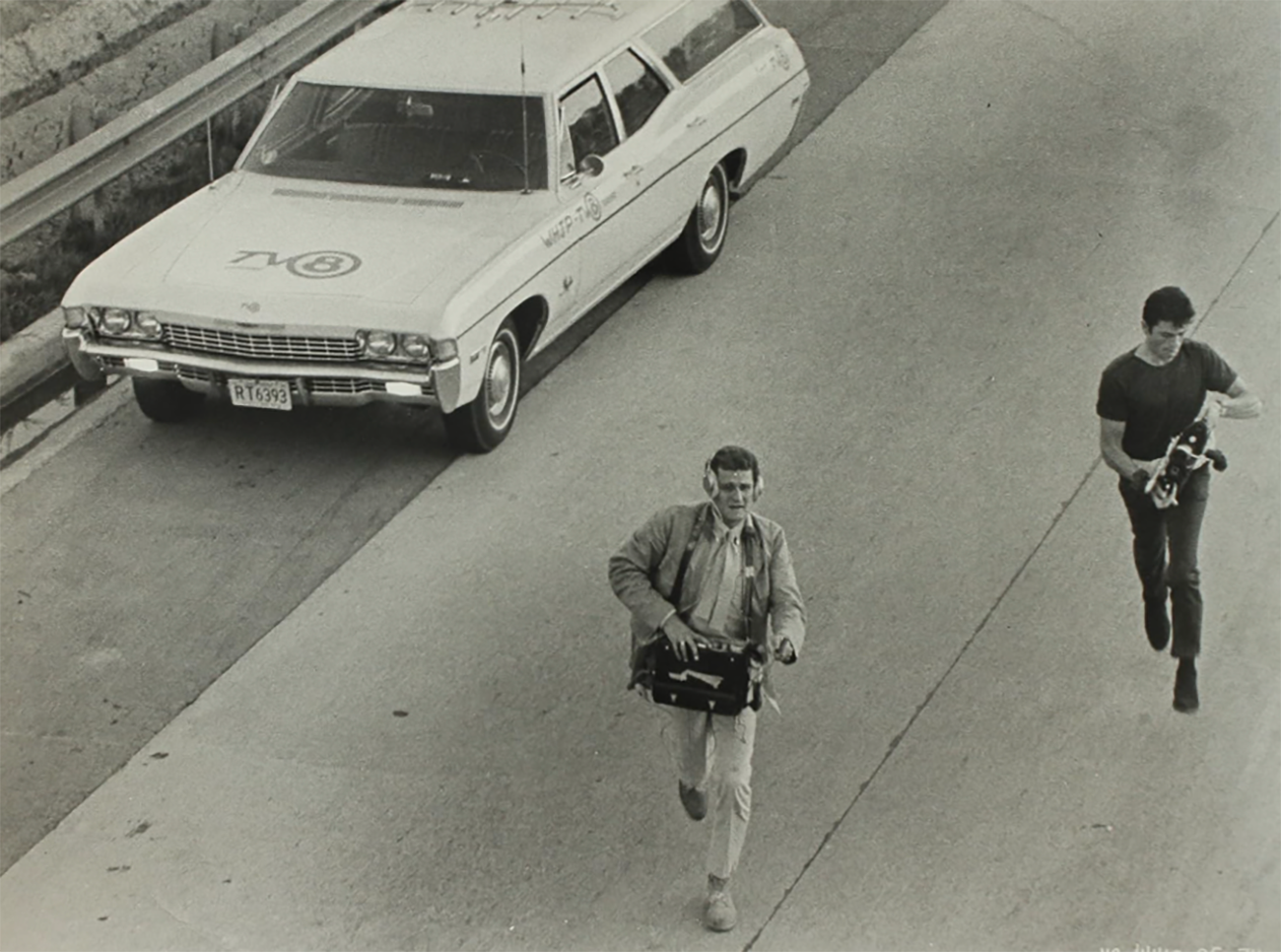 Peter Bonerz and Robert Forster carrying camera equipment in a film still from the 1960s.