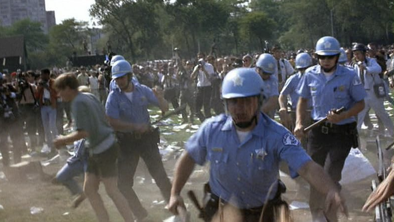 “Real” footage from the 1968 Chicago protests show a crowd of police and protesters clashing on a grassy lawn strewn with papers.