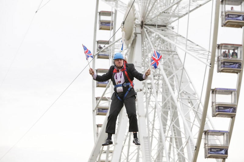 A main in a suit and helmet holds a British flags in each hand as he hangs from a zipline in front of a ferris wheel.