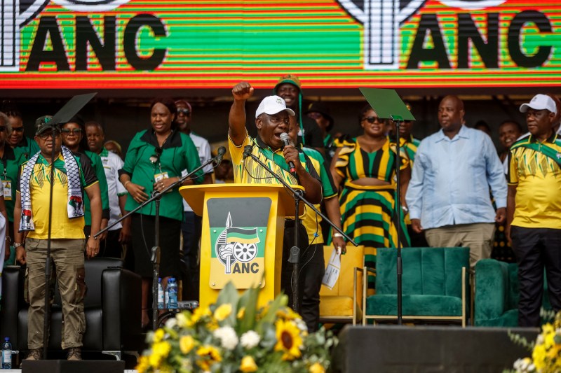 South African President Cyril Ramaphosa speaks at a podium onstage at an ANC rally. Zuma wears a shirt in the green-and-yellow party colors, as do many of the people crowded onstage behind him. He also raises one fist in the air as he speaks into the microphone.