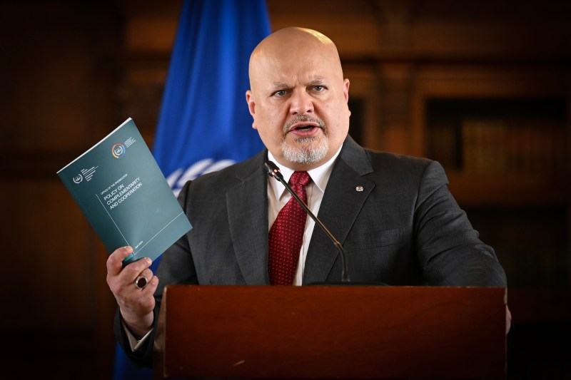 The International Criminal Court’s top prosecutor, Karim Khan, a bald man in a gray suit and red tie, speaks at a podium during a press conference in Bogota. A blue ICC flag hangs behind him in front of a wood-paneled wall.
