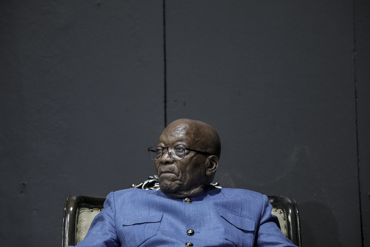 Zuma is seen from the chest upward, wearing a blue jacket buttoned to his neck as he sits in a deep chair against a dark gray wall. He wears a serious expression as he looks slightly to the left.