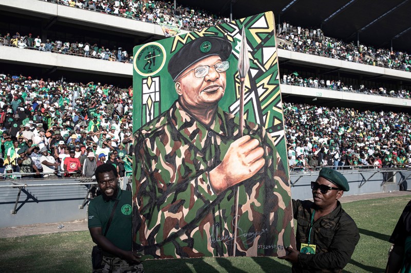 Two men, one in a t-shirt, the other in a uniform, beret and sunglasses, hold up a large painted portrait of Jacob Zuma in military garb and holding a spear. The men stand in front of a large stadium, with the crowd seen behind them.