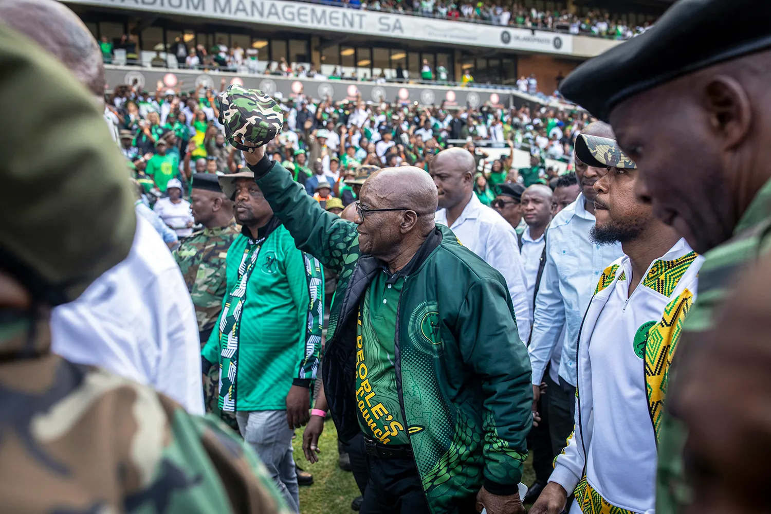 Jacob Zuma, wearing a green bomber jacket and a green-and-yellow campaign shirt, raises his hand to wave as he's surrounded by staff and security members. A large stadium crowd is seen in the stands in the background, many of them dressed in green and yellow.