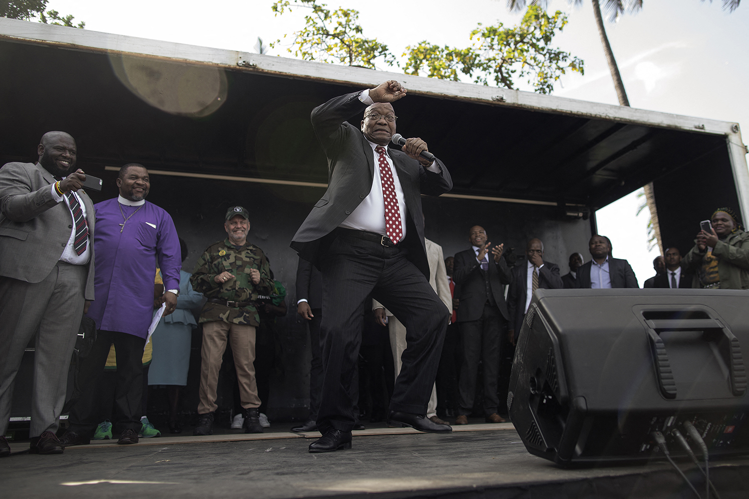 Zuma, wearing a suit and tie and holding a microphone, sings and dances with one hand raised in the air. Behind him, several others smile and clap onstage.