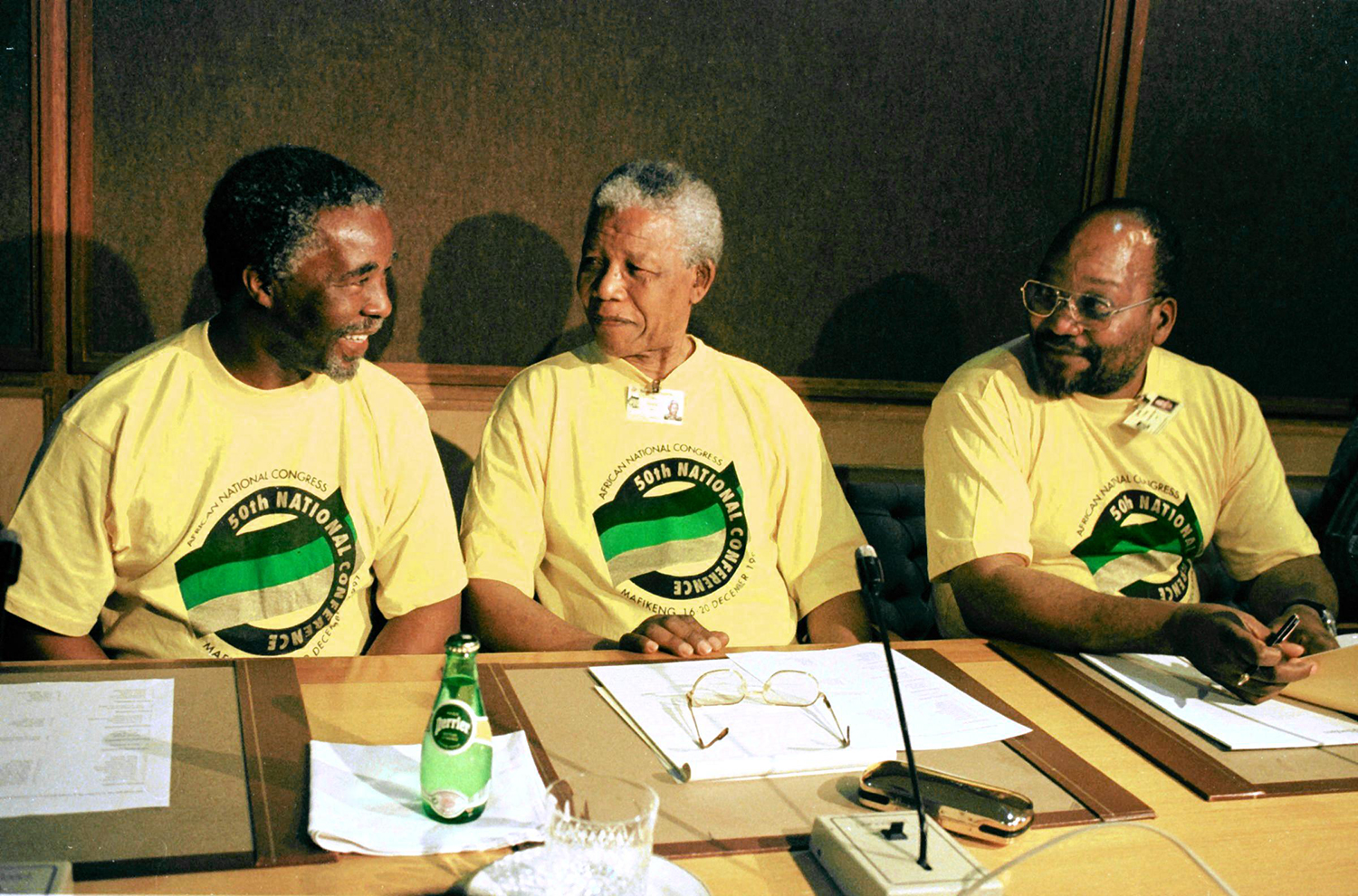 Three men wearing 50th National Conference ANC T-shirts sit behind a table that holds a microphone, glasses, papers, a Perrier bottle and water glass.