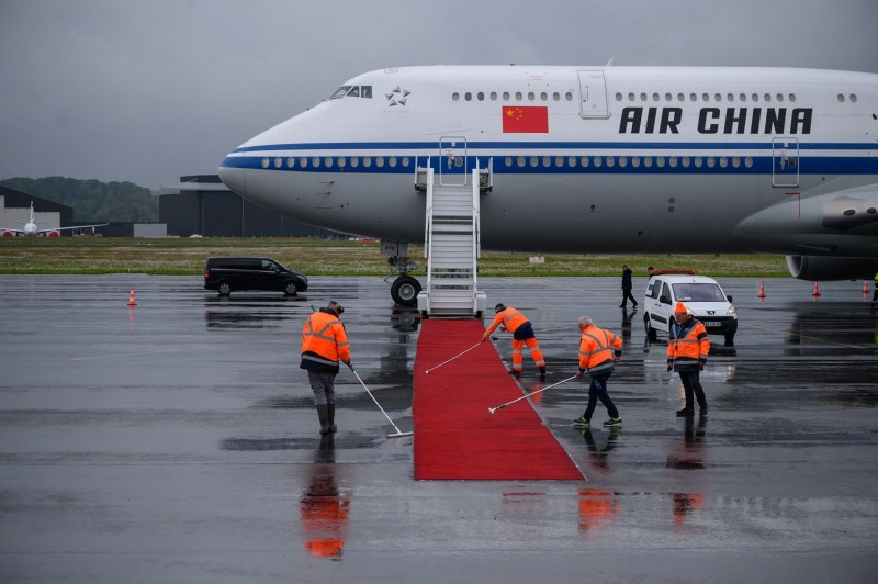 Workers wearing reflective orange jackets stand on an airport tarmac and sweep a red carpet that extends from the boarding steps of the Chinese president's airplane. The pavement around the carpet is damp with puddles, and the sky overhead is gray and overcast.