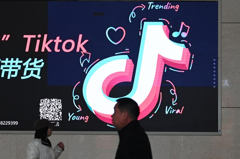 Two people walk past a sign for TikTok on a wall. Scribbles on the sign annotate the TikTok logo with the words "young," "trending," and "viral" in English along with characters in Chinese.