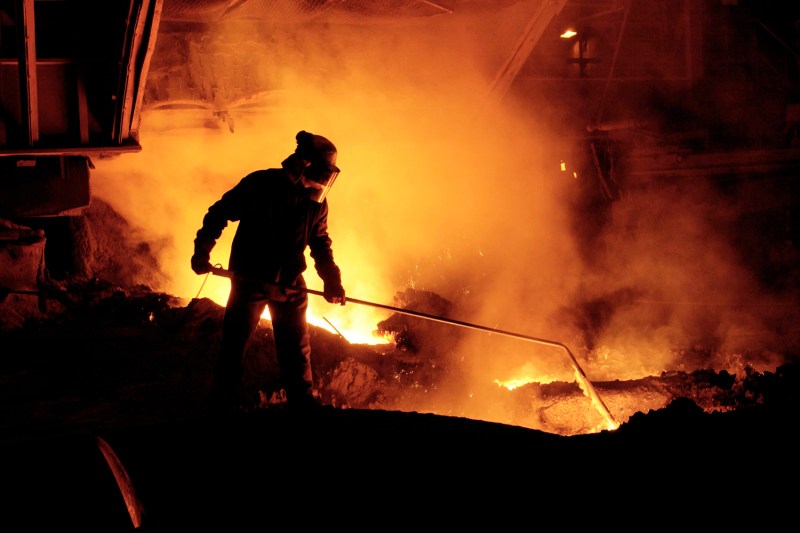 A steel worker takes an iron sample from a blast furnace, surrounded by molten iron.