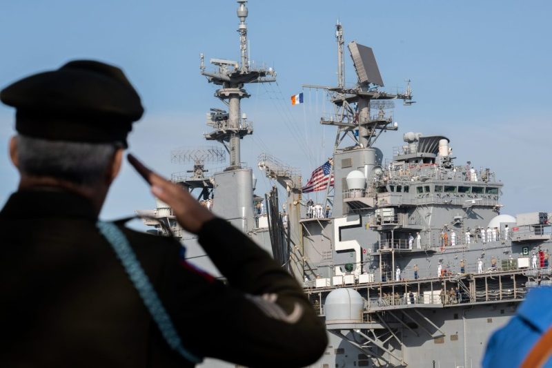 A uniformed person is seen from behind saluting a large navy ship with sailors on the deck.