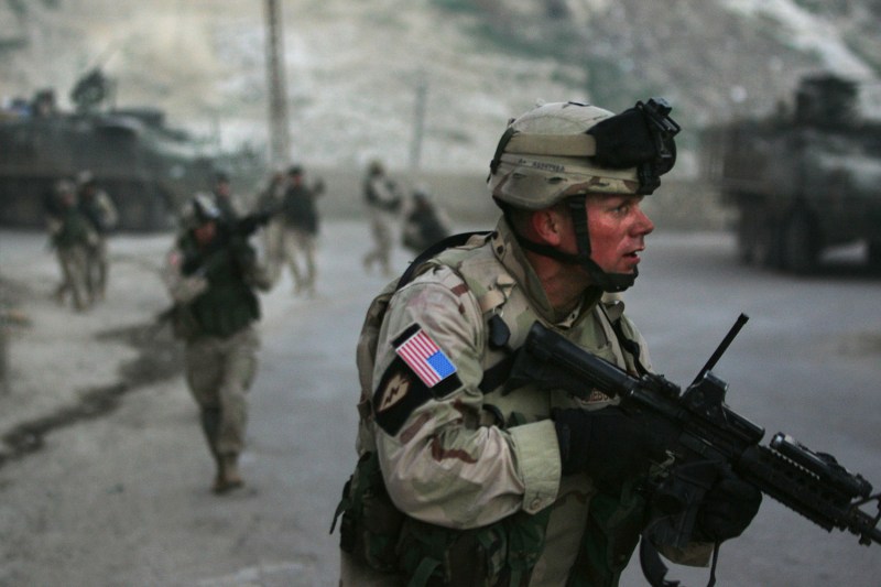 A U.S. solderi holding a gun and wearing a helmet provides cover for other soldiers seen behind him out of focus.