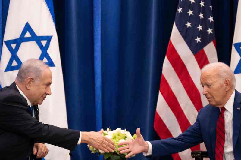 U.S. President Joe Biden reaches his arm out a table to shake hands with Israeli Prime Minister Benjamin Netanyahu. Both men wear dark suits and ties, and the Israeli and American flags hang in front of a blue curtain beyond the two leaders.