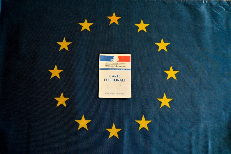 A French electoral card is laid atop a European Union flag, centered between the circle of yellow stars against the dark blue background. The card is labeled with French text and also shows the colors of the French flag and the national motto: "Liberty, equality, fraternity, or death."