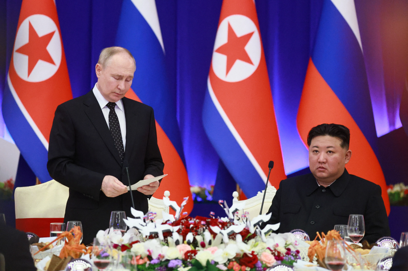 Russian President Vladimir Putin (left) stands at a table set with food and flowers and reads aloud from a sheet of paper as North Korean leader Kim Jong Un, seated, listens to him. The Russian and North Korean flags hang at the back of the room.