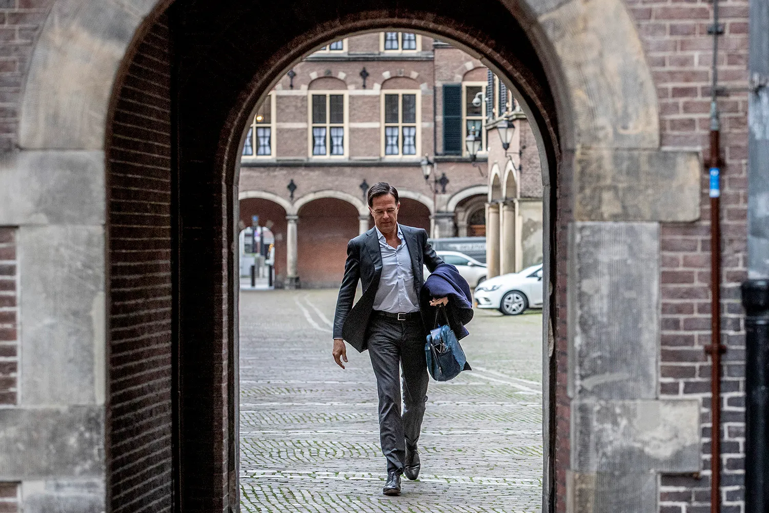 Rutte, wearing an open suit jacket, button-up shirt, and no tie, carries a bag in one hand as he walks through an arched stone entrance. The cobblestone courtyard that Rutte just crossed is visible through the doorway.