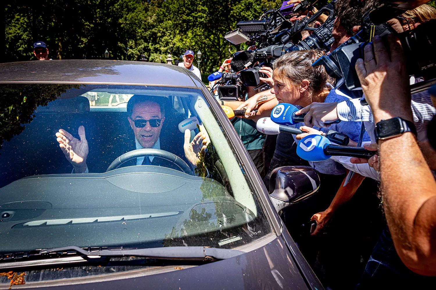 Mark Rutte gestures with both hands, his mouth open, as he sits behind the wheel of a car as reporters with microphones and video cameras swarm the vehicle, some of them sticking microphones through the open drivers' side window. Rutte wears a dark suit and tie with sunglasses.
