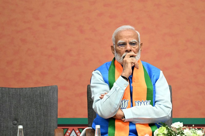 Indian Prime Minister Narendra Modi sits onstage at a party meeting. He crosses his arms and puts one hand over his face in contemplation as he watches the proceedings.
