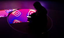 A person sitting in a dark room on their phone. A pattern showing the Instagram logo is reflected in mirrors below them. 
