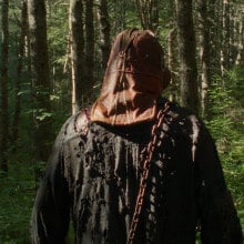 A man wearing a mask is seen from behind as he walks through the woods.