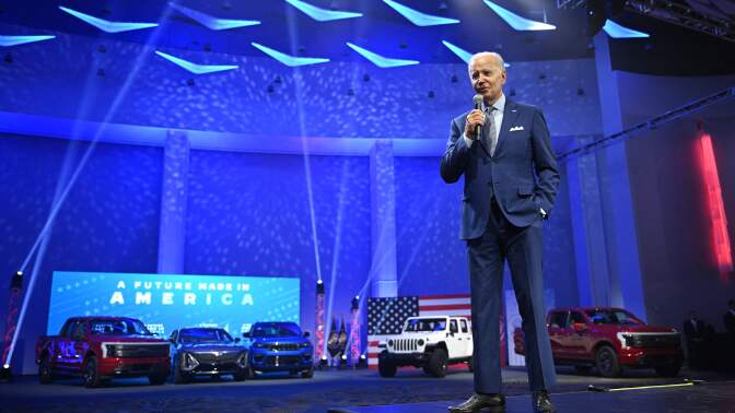 Old white man in blue suit speaks into a microphone. In the background is a display lined with electric vehicles in red, white, and blue variations