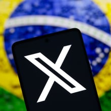 The X (Twitter) logo is displayed on a smartphone screen with a flag of Brazil in the background. Social network X has announced that it will close its office in Brazil.