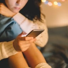 A girl dressed in a white sweater looks at her phone, while sitting in her bedroom.