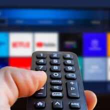 a right hand holds a remote control pointed at a TV with streaming app icons shown on the screen