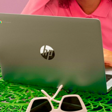 a woman working on a HP Chromebook 14a-ne0013dx at a green table that has a cup of tea and a pair of sunglasses sitting on it