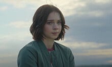 A teenage girl stands in front of a blue sky backdrop, looking worried.