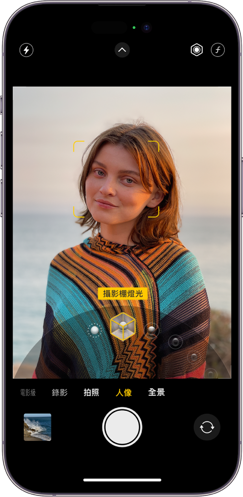處於「人像」模式的「相機」畫面，在觀景窗中，主體很清晰且背景已模糊化。觀景窗底部打開用來選取「人像燈光」的轉盤，選取了「攝影棚燈光」。畫面左上角為「閃光燈」按鈕，頂端中央為「相機控制項目」按鈕，右上角的按鈕用來調整「人像燈光」強度和「景深控制」。螢幕底部由左至右為「照片和影片檢視器」按鈕、「拍照」按鈕和「相機選擇器後置」按鈕。