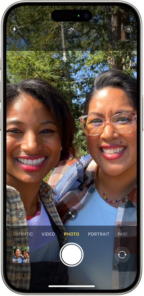 La pantalla de la app Cámara en el modo Foto. Debajo de los modos de cámara, de izquierda a derecha, se encuentran los botones Visor de fotos y videos, Tomar foto y Selector de cámara posterior. En la parte superior de la pantalla se encuentran los botones Flash, Controles de la cámara y Live Photos. Dentro del marco de la cámara, las personas se están tomando una selfie.